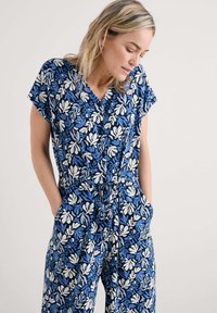 Niebieski kwiatowy kombinezon z krótkimi rękawami, dekoltem w kształcie V, wiązaniem w talii i kieszeniami; wzór zawiera białe i jasnoniebieskie liście na ciemnym tle.