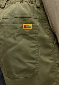 Olívazöld nadrág nagy hátsó zsebbel és a zseb fölött varrt sárga Fjällräven logós címkével.