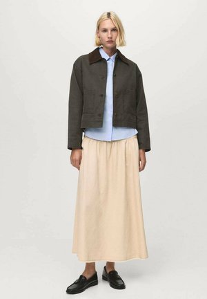 Giacca verde scuro tagliata con colletto marrone, camicia azzurro chiaro e gonna lunga beige. Mocassini neri completano l'outfit.