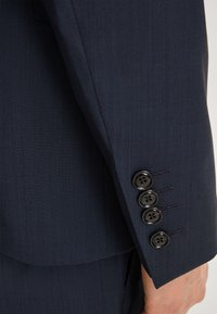 Dopasowana marynarka w kolorze granatowym, z rękawem wykonanym z teksturowanej tkaniny, posiadająca pięć czarnych guzików oraz widoczne detale szycia w okolicy mankietu.