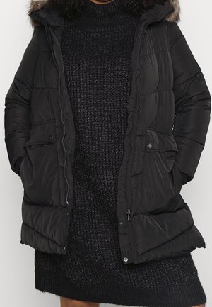 Osoba nosící černou prošívanou péřovou bundu s kožešinou lemovanou kapucí přes černé pletené šaty, ruce v kapsách bundy.