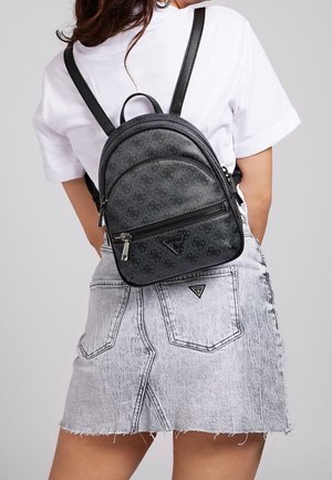 Vrouw in een zwart patroon rugzak over een wit t-shirt en een acid-wash spijkerrok met achterzakken en een klein logo patch.