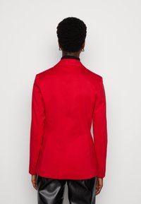Czerwona marynarka o dopasowanym kroju z wyraźną strukturą, z klapami w kształcie notcha, tyłem z jednym rozcięciem i zapinanymi mankietami, zestawiona z czarnymi spodniami.