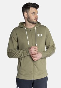 Oliwkowy rozpinany hoodie z przednią kieszenią, białymi sznurkami i białym logo Under Armour na klatce piersiowej. Miękka, teksturowana tkanina.