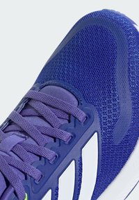 Chaussure de sport violette avec empeigne en maille, accents texturés et lacets plats. Présente trois rayures blanches et un détail de talon dégradé.