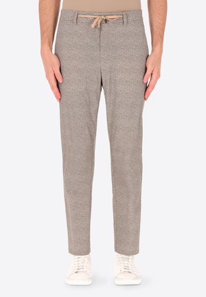 Uomo che indossa pantaloni beige con coulisse e texture, e scarpe da ginnastica bianche, in piedi con le braccia rilassate lungo i fianchi su uno sfondo chiaro e uniforme.