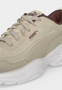 Μπεζ αθλητικό παπούτσι Puma με καφέ λογότυπο, μπεζ κορδόνια και χοντρή λευκή σόλα, που δείχνει λεπτομέρειες από το πλάι και επάνω.