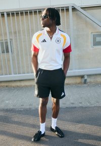 Hvit polo skjorte med røde og gule detaljer, svarte shorts og svarte sko. Har Adidas-merke og Tysklands flagg-emblem.