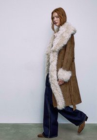 Brun ruskind frakke med en fyldig hvit faux fur krave og manchetter, lange ærmer og sidelommer, parret med brede denimbukser.