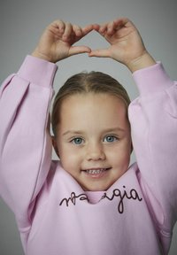 Ragazza giovane con occhi blu che indossa un felpa rosa, forma un cuore con le mani sopra la testa, sorridendo verso la fotocamera.