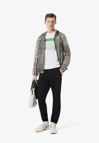 Joven vistiendo una chaqueta con patrón de cuadrícula negro y beige, camiseta blanca con el logo de Lacoste, pantalones negros, zapatillas blancas, sosteniendo ropa doblada con la mano izquierda.
