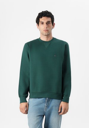 Green sweatshirt with crew neck, ribbed cuffs, and hem. Features a small logo on the chest. Paired with light blue jeans.