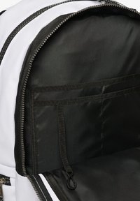 Intérieur noir d'un sac à dos avec une fermeture éclair, comportant deux poches élastiques et une boucle suspendue pour les clés ou les accessoires. Extérieur blanc et accents noirs.