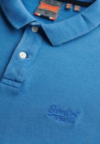 Polo bleu avec un tissu texturé, trois boutons blancs et un logo brodé dans le coin inférieur. Étiquette d'entretien visible dans l'encolure.