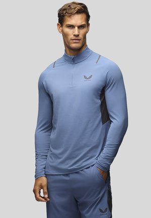 Homme portant un haut de sport bleu clair à manches longues avec fermeture éclair quart de zip et un short assorti, debout avec une main dans la poche sur un fond uni.