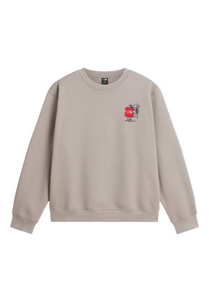 Persona que lleva una sudadera beige con gráfico de un excursionista que lleva equipo de escalada, cuerdas y herramientas en la espalda, combinado con una gorra clara.