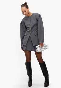 Blazer gris à manches surdimensionnées, boutons argentés, jupe plissée assortie, bottes noires montantes jusqu'aux genoux, et pochette argentée cloutée.