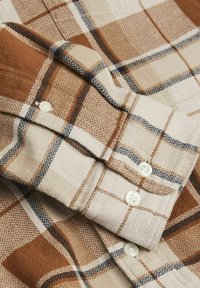 Rjava in bež karirasta srajca s gumbi, mehke teksture; vsebuje bele gumbe, kontrastne modre črte in zložljiv ovratnik.