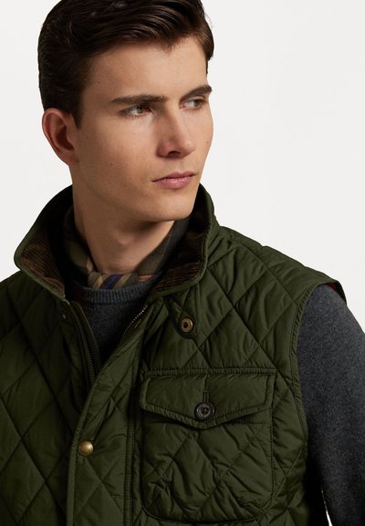 Zaļa quilted veste ar augstu apkakli un pogu aizdari. Iespējo krūšpogu un mīkstu ruļļu oderi, uzvilkta virs pelēka džempera.