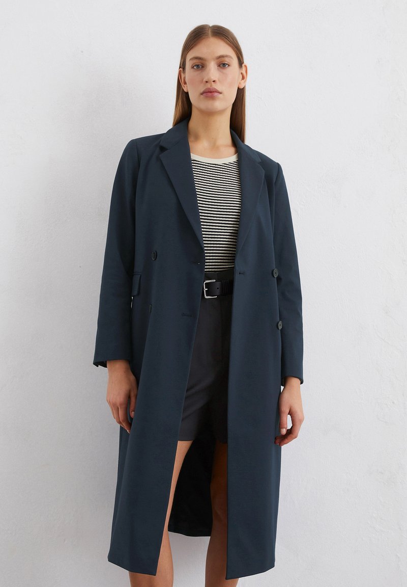 Dunkelblauer, doppelt-knöpfbarer Trenchcoat mit Revers, langen Ärmeln und geradem Schnitt, kombiniert mit einem gestreiften Oberteil und Shorts.