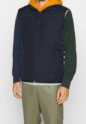 Mężczyzna w granatowej pikowanej kamizelce na kolorowej bluzie z kapturem — kaptur pomarańczowy, prawy rękaw zielony, lewy niebieski, w połączeniu z beżowymi spodniami.