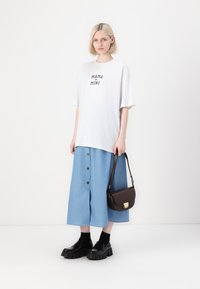 T-shirt blanc avec le texte "MAMA + MINI", associé à une jupe légère bleu clair à boutons sur le devant. Des bottes à plateforme noires et un sac bandoulière marron complètent le look.