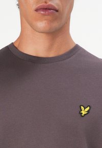 Mann, der ein einfarbiges lila Rundhals-Shirt trägt, mit einem kleinen gelben Adler-Logo, das auf der linken Brust gestickt ist.