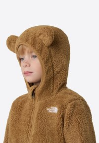 Rjava flis jopa s kapuco z medvedjimi ušesi, teksturirano tkanino, polnim zadrgo in logotipom na prsih; ima tesno, okroglo kapuco.