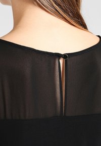 Mustapaita, jossa on läpinäkyvä kangasosuus ylhäällä, pyöreä pääntie ja pieni avaus takana, joka on kiinnitetty mustalla napilla.