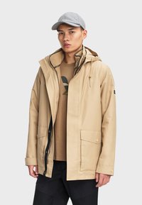 Beige wasserdichte Jacke mit Reißverschluss und Druckknopfverschluss, Kapuze, großen frontalen Taschen und strukturiertem Innenfutter. Kombiniert mit einem braunen Hemd und einer Mütze.