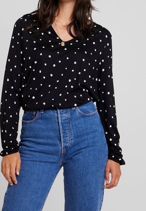 Frau trägt eine schwarze Bluse mit langen Ärmeln und weißen Punkten, die in eine hoch taillierte blaue Jeans gesteckt ist, vor einem schlichten Hintergrund.