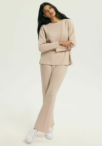 Beżowy komplet dzianinowy do spędzania czasu w domu, składający się z długiego topu z okrągłym dekoltem i rozszerzanych spodni, zestawiony z białymi trampkami. Miękka tekstura i luźny krój.