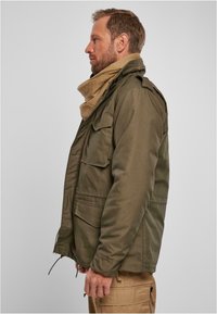 Chaqueta verde oliva con múltiples bolsillos frontales, epaulettes en los hombros y dobladillo con cordón. Combinada con una bufanda beige alrededor del cuello.