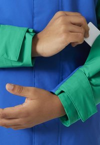 Niebieska i zielona kurtka charakteryzuje się eleganckim designem z kontrastującymi mankietami. Gładka tkanina z widocznym przeszyciem i płaskim kołnierzykiem. Ręce regulujące rękaw.