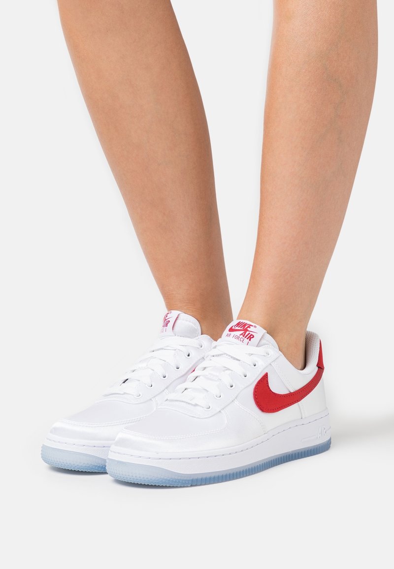 Baskets blanches Nike Air Force 1 avec le swoosh rouge et le logo, portées aux pieds sur un fond blanc uni.