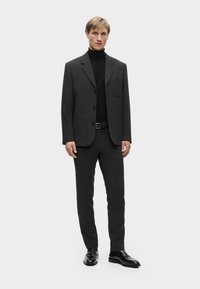 Abito grigio scuro con blazer aderente, rever a punta e tasche frontali, abbinato a un dolcevita nero e mocassini neri lucidi.