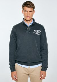 Tumehall pullover-sviiter, millel on valekaelus ja rindade graafika valges. Pehme tekstuur, ribikud mansetid ja äär. Kanti helesinise särgi peal.