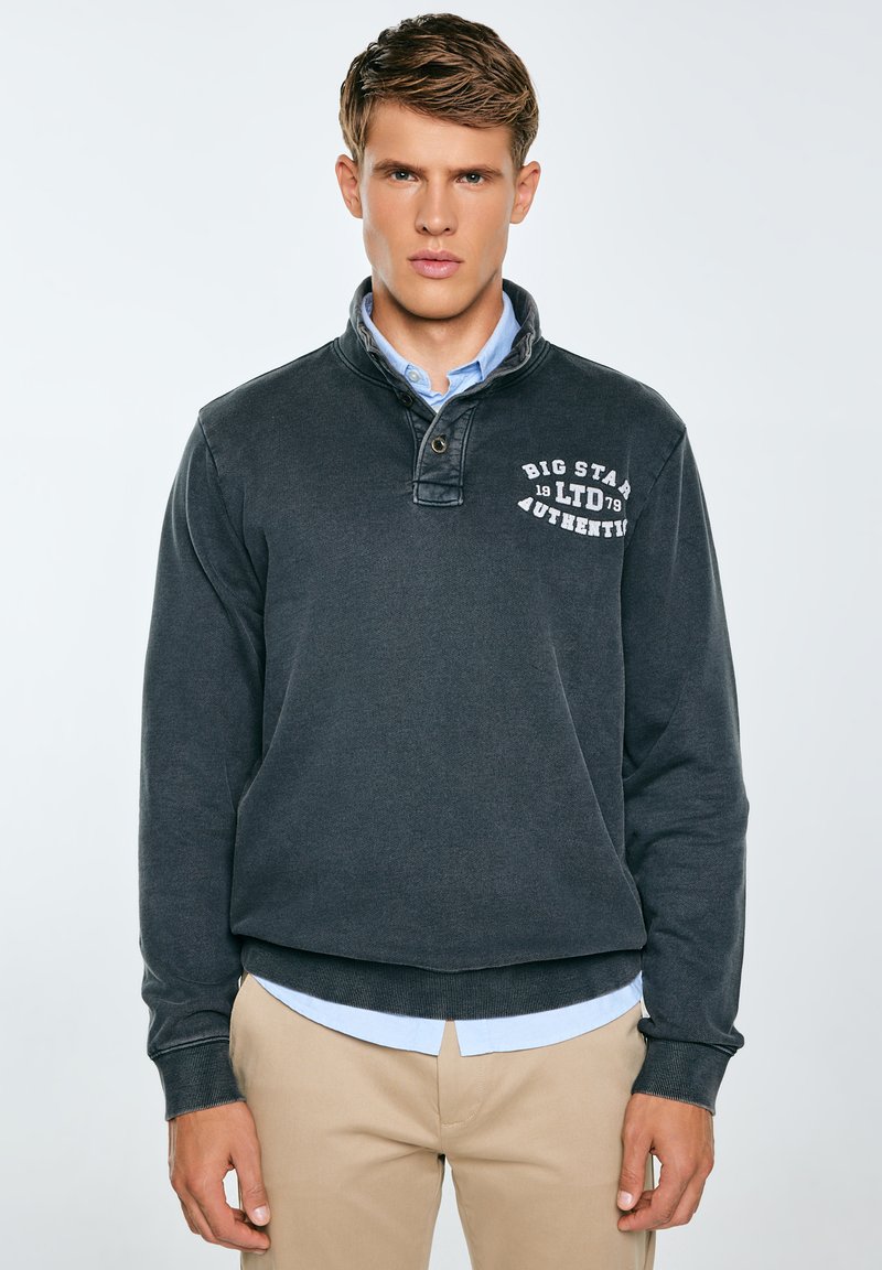 Tumehall pullover-sviiter, millel on valekaelus ja rindade graafika valges. Pehme tekstuur, ribikud mansetid ja äär. Kanti helesinise särgi peal.