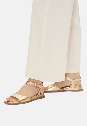 Nohy obuté v metalicky zlatých sandálech s otevřenou špičkou, plochou podrážkou, pásky kolem kotníků a přezkami, v kombinaci s bílými širokými kalhotami.