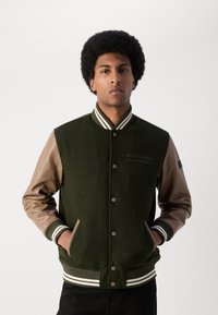 Chaqueta varsity de mezcla de lana verde con mangas de cuero marrón, cuello y puños de canalé a rayas, botones a presión y bolsillos laterales.
