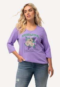Camiseta de algodón morada con mangas tres cuartos, que presenta un gráfico floral y el texto "Sociedad HORTICULTURAL". Textura suave y corte relajado.
