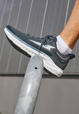 Person, die einen schwarzen Laufschuh trägt und über ein silbernes Metallgeländer tritt, mit grauer Wand im Hintergrund.