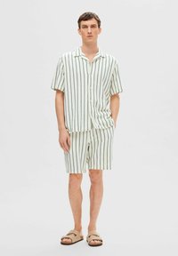 Šviesiai žalia trumpomis rankovėmis marškinėliai su mygtukais ir atitinkančios šortai, dekoruoti tamsiomis vertikaliomis juostelėmis, pagaminti iš tekstūruoto audinio. Avimi kasdieniai sandalai.