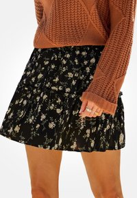 Černá květinová sukně s vrstevnatým volánkovým designem, zdobená malými bílými a zelenými květinami. Nosit s texturovaným pleteným svetrem v barvě rzi.