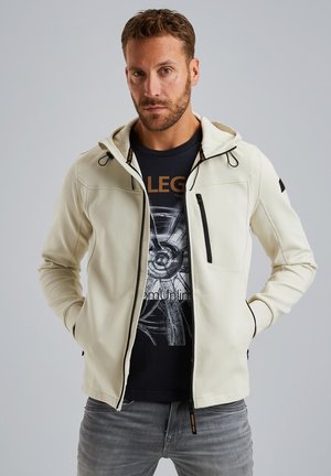 Light beige hooded jacket with black accents, zippered pockets, and a soft, smooth texture. Worn over a black graphic t-shirt and gray jeans.