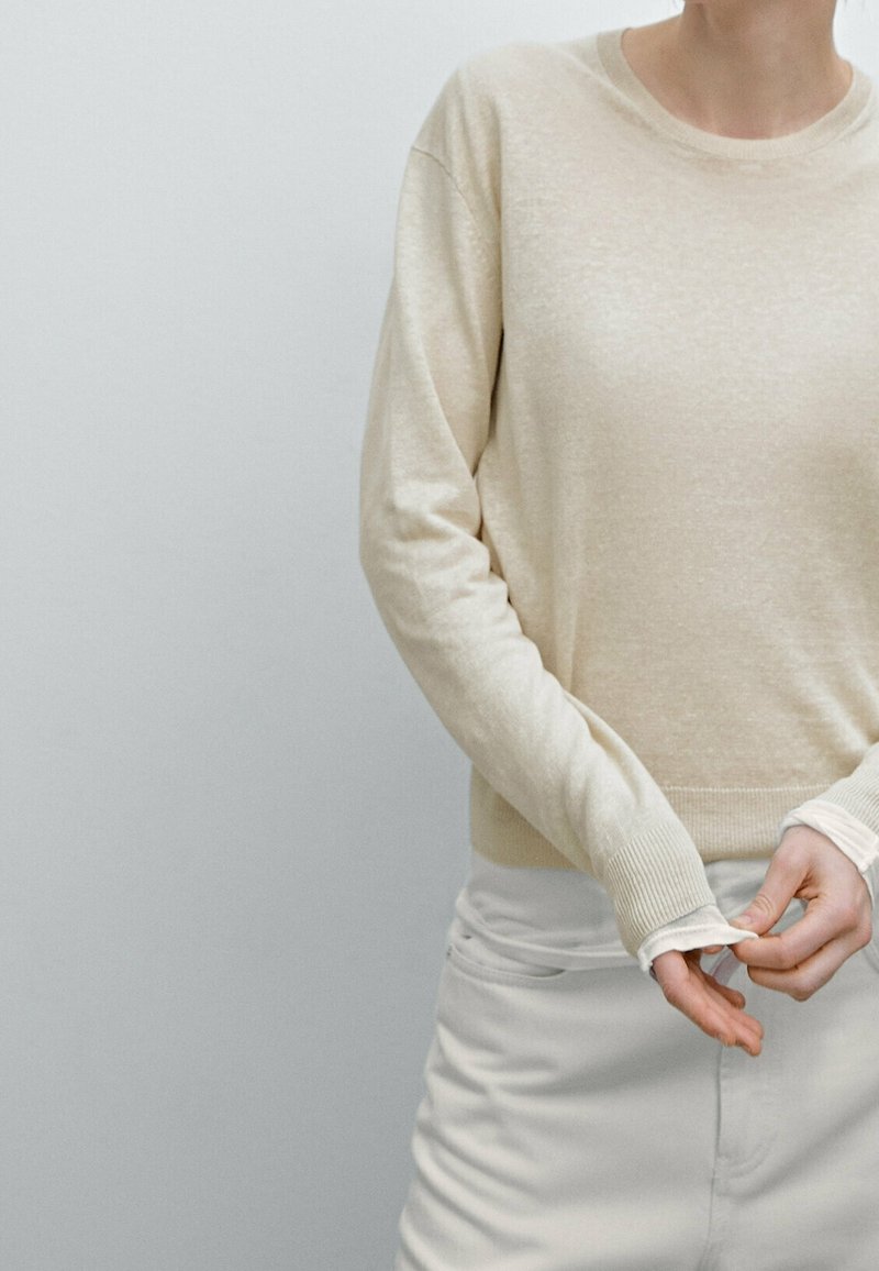 Personne portant un pull beige et un pantalon blanc ajustant sa manche devant un fond gris clair uni.