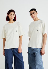 Krémové trička s krátkým rukávem a kapsou na hrudi; jedno z nich má malý, barevně vyšívaný design. V kombinaci s širokými džíny.
