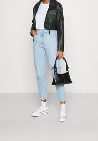 Person som bär en svart läderjacka, vit t-shirt, ljusblå rivna jeans, vita hög-top sneakers och håller en svart handväska.