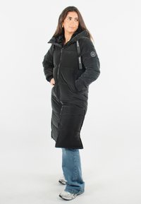 Doudoune noire avec capuche, longue, dotée d'une fermeture éclair et d'un patch logo sur le bras ; texture lisse avec un motif de coutures en relief.