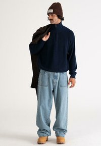 Man wearing brown knit beanie, navy half-zip sweater, light blue wide jeans, tan boots, holding dark coat over shoulder, standing on white background.